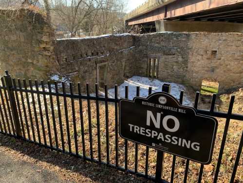 Ruins of Simpsonville Mill behind a black fence with a "No Trespassing" sign, under a bridge and surrounded by snow.