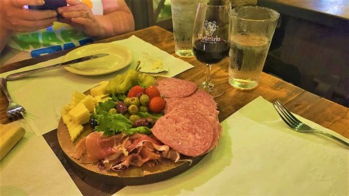 A wooden platter with assorted meats, cheeses, olives, and cherry tomatoes, accompanied by drinks and a person using a phone.