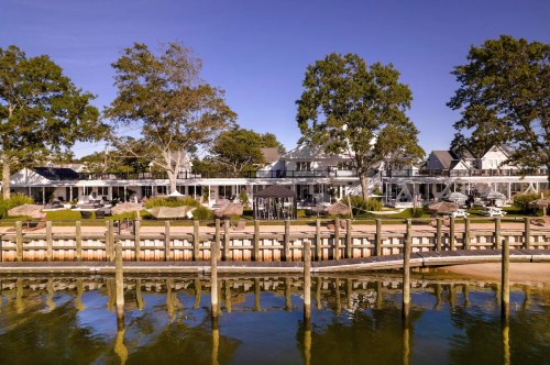 A serene waterfront view of a resort with trees, a dock, and calm water under a clear blue sky.