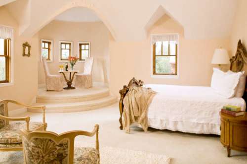 Cozy bedroom with a bed, elegant chairs, and a small dining area by the windows, featuring soft, warm colors.