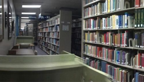 A view of a library aisle lined with bookshelves, featuring tables and chairs in the foreground.