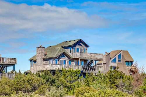 A large, multi-level beach house with a green roof, surrounded by trees under a blue sky.