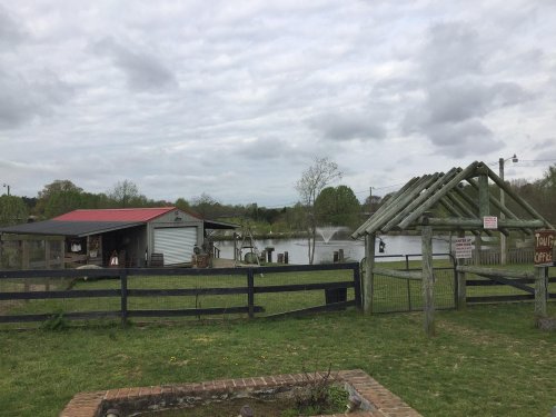 A rustic scene featuring a pond, a barn with a red roof, and a wooden shelter under a cloudy sky.