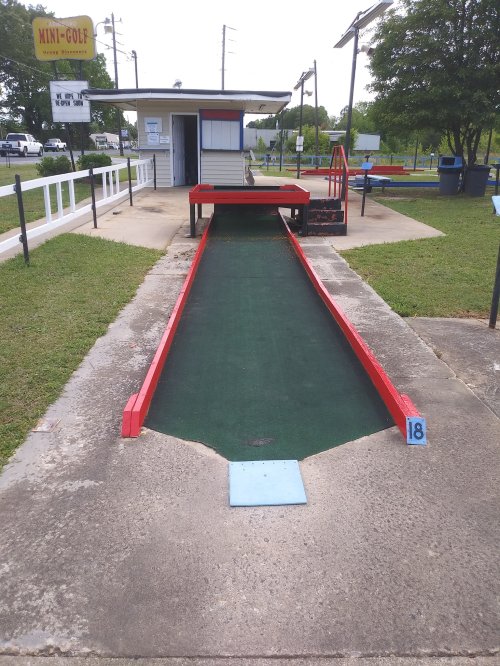 A mini-golf course with a green carpeted hole, red borders, and a small building in the background.