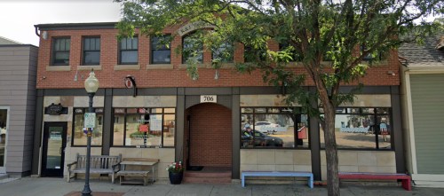 A brick building with large windows, benches outside, and a sign above the entrance, located on a street.