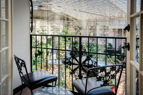 A balcony with two black chairs overlooks a lush indoor garden with a glass ceiling and decorative lights.