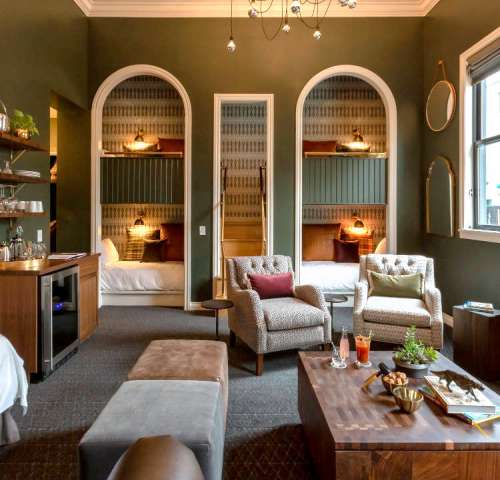 Cozy hotel room with green walls, two beds in alcoves, stylish seating, and a small bar area.