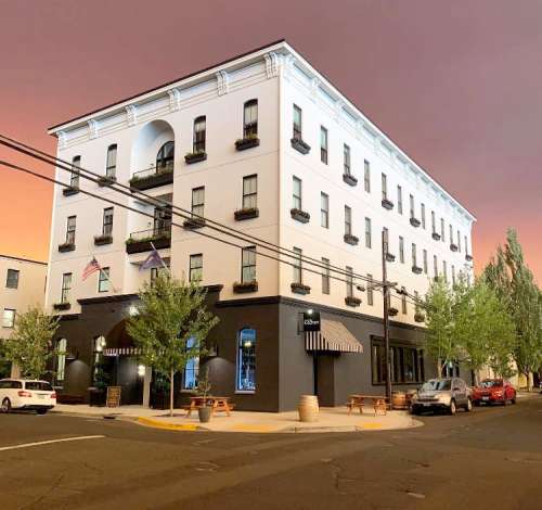 A modern three-story hotel with balconies, surrounded by trees, under a colorful sunset sky.