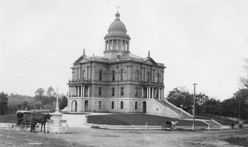 Historic black-and-white photo of a grand building with a dome, surrounded by trees and horse-drawn carriages.