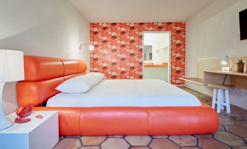 A modern hotel room featuring an orange bed, patterned wall, desk, and a cozy atmosphere.