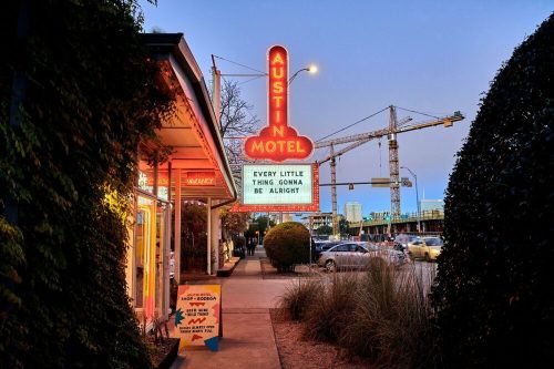 Neon sign of Austin Motel at dusk, with a message: "Every little thing gonna be alright," surrounded by greenery and traffic.