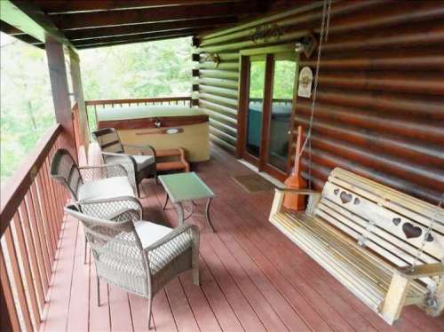 A cozy porch with a swing, chairs, and a hot tub, surrounded by greenery and wooden logs.