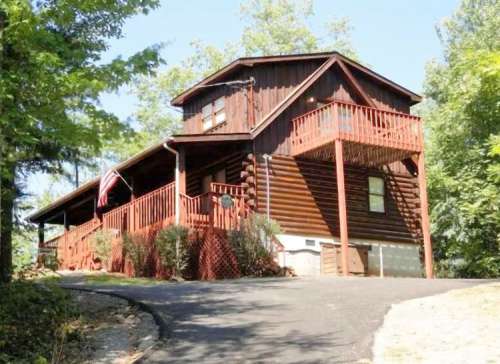 A rustic log cabin with a red balcony, surrounded by trees and a paved driveway, featuring an American flag.