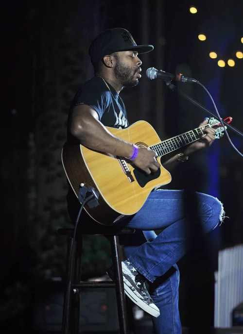 A musician plays acoustic guitar and sings into a microphone on stage, illuminated by soft lights.