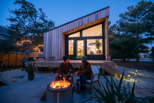 A cozy outdoor scene with two people by a fire pit, enjoying the evening near a modern wooden house.