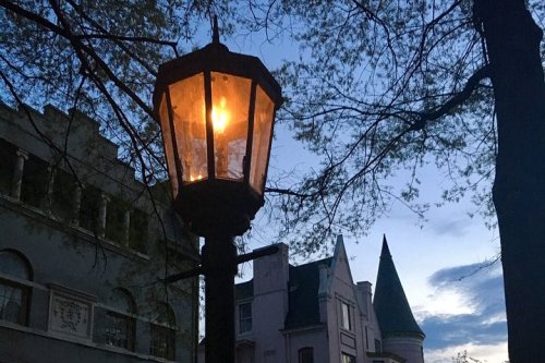 A glowing lantern stands on a post, with trees and buildings in the background under a twilight sky.