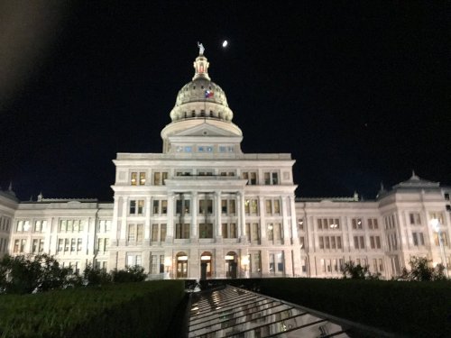 The Texas State Capitol building illuminated at night, with a crescent moon visible in the sky above.