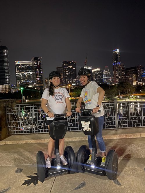 Two women on segways at night, smiling with a city skyline in the background, wearing helmets and casual clothing.