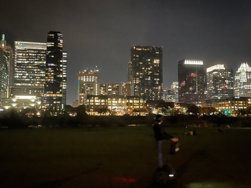 City skyline at night with illuminated buildings and a person in the foreground holding a bag.