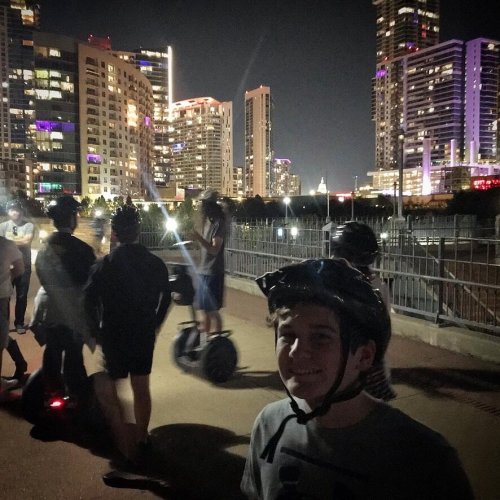 A group of people riding scooters and segways at night, with city skyscrapers illuminated in the background.