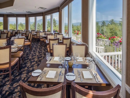 Elegant restaurant interior with tables set for dining, large windows showcasing a scenic view and floral decorations.