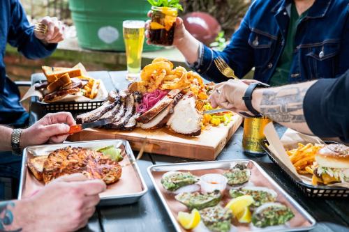 A table filled with various dishes, including grilled meats, fries, onion rings, and drinks, with hands reaching for food.