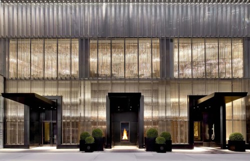 Modern hotel entrance with sleek glass facade, illuminated by warm lights and flanked by neatly trimmed greenery.