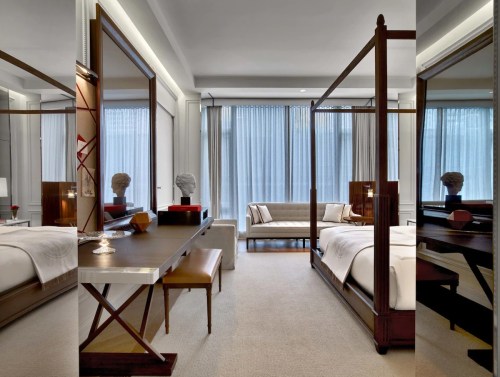 A modern hotel room featuring a four-poster bed, a desk, a sofa, and large windows with sheer curtains.