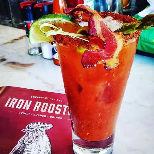 A vibrant cocktail garnished with lime, bacon, and spices, next to a menu titled "Iron Rooster."
