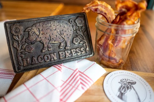 A bacon press next to a jar of crispy bacon, with a napkin and a coaster on a wooden table.