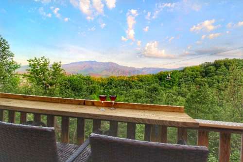 Two glasses of red wine sit on a wooden railing, overlooking a lush green landscape and distant mountains under a blue sky.