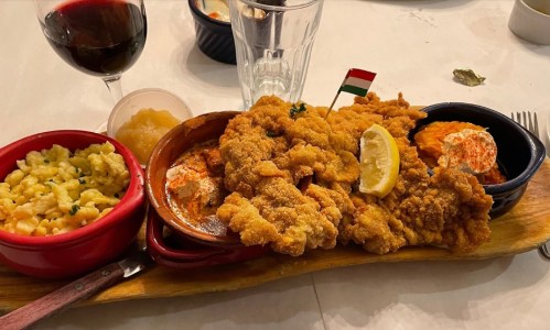 A wooden platter with fried chicken, sides of macaroni, spicy sauce, and a lemon wedge, accompanied by a glass of wine.
