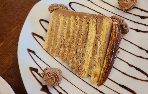 A slice of layered chocolate cake on a white plate, drizzled with chocolate sauce and decorated with chocolate swirls.