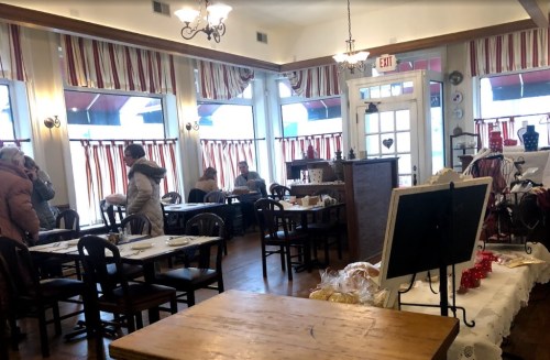 A cozy restaurant interior with wooden tables, chairs, and large windows, featuring diners and decorative elements.