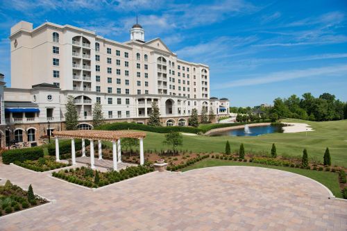 A luxurious hotel with a manicured lawn, pond, and blue sky, featuring a pergola in the foreground.