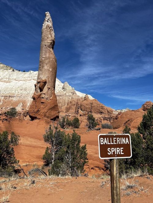 A tall rock formation known as Ballerina Spire stands against a blue sky, surrounded by red rock and green shrubs.