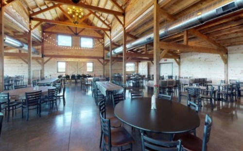 A spacious wooden barn interior with round tables, chairs, and string lights, ready for an event.
