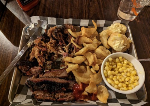 A metal tray with pulled pork, brisket, fried snacks, and a bowl of corn, served in a casual dining setting.