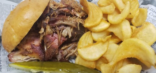 A pulled pork sandwich with crispy potato chips and a pickle slice, served on a tray lined with newspaper.
