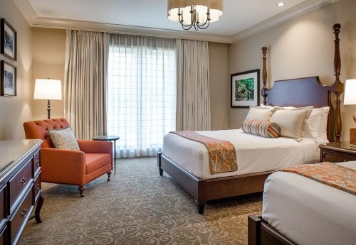 Cozy hotel room with two beds, an orange armchair, dresser, and large windows with sheer curtains.