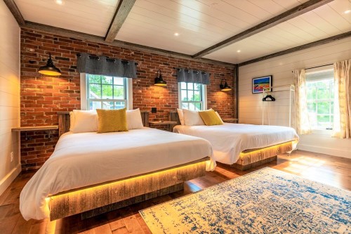 Cozy bedroom with two beds, exposed brick walls, wooden beams, and large windows letting in natural light.