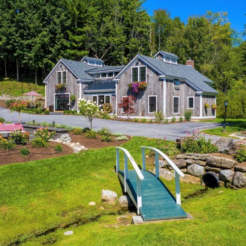 A charming wooden house surrounded by greenery, flowers, and a small bridge over a stream in a sunny landscape.