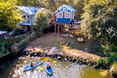 Aerial view of a blue house on stilts by a river, with two people kayaking nearby. Lush greenery surrounds the area.