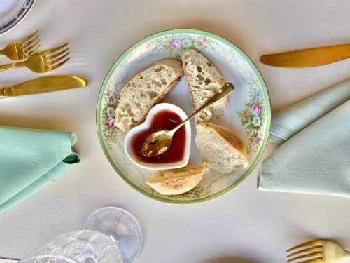 A heart-shaped dish with jam sits on a floral plate, surrounded by bread slices and gold utensils.