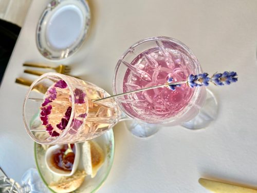 Two elegant glasses with pink drinks, garnished with lavender and edible flowers, on a table with light snacks.