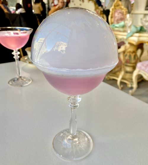 A pink cocktail in a glass topped with a dome of fog, set on a white table with blurred background.