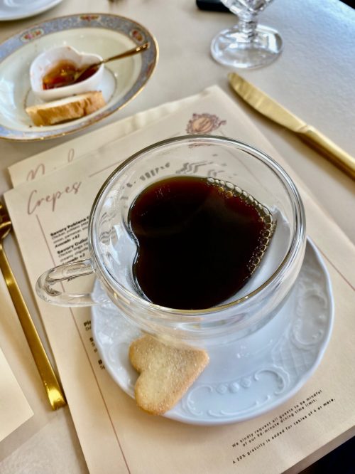 A heart-shaped cup of coffee sits on a saucer with a small heart-shaped cookie beside it, on a table with menus.