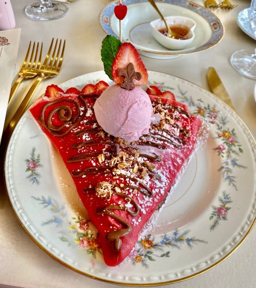 A colorful dessert featuring a strawberry crepe topped with chocolate drizzle, whipped cream, and a scoop of pink ice cream.