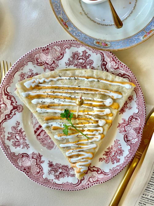 A triangular crepe drizzled with sauce on a decorative plate, garnished with a small herb.