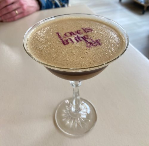 A cocktail in a coupe glass with foam on top, featuring the words "Love is in the Air" written in pink.